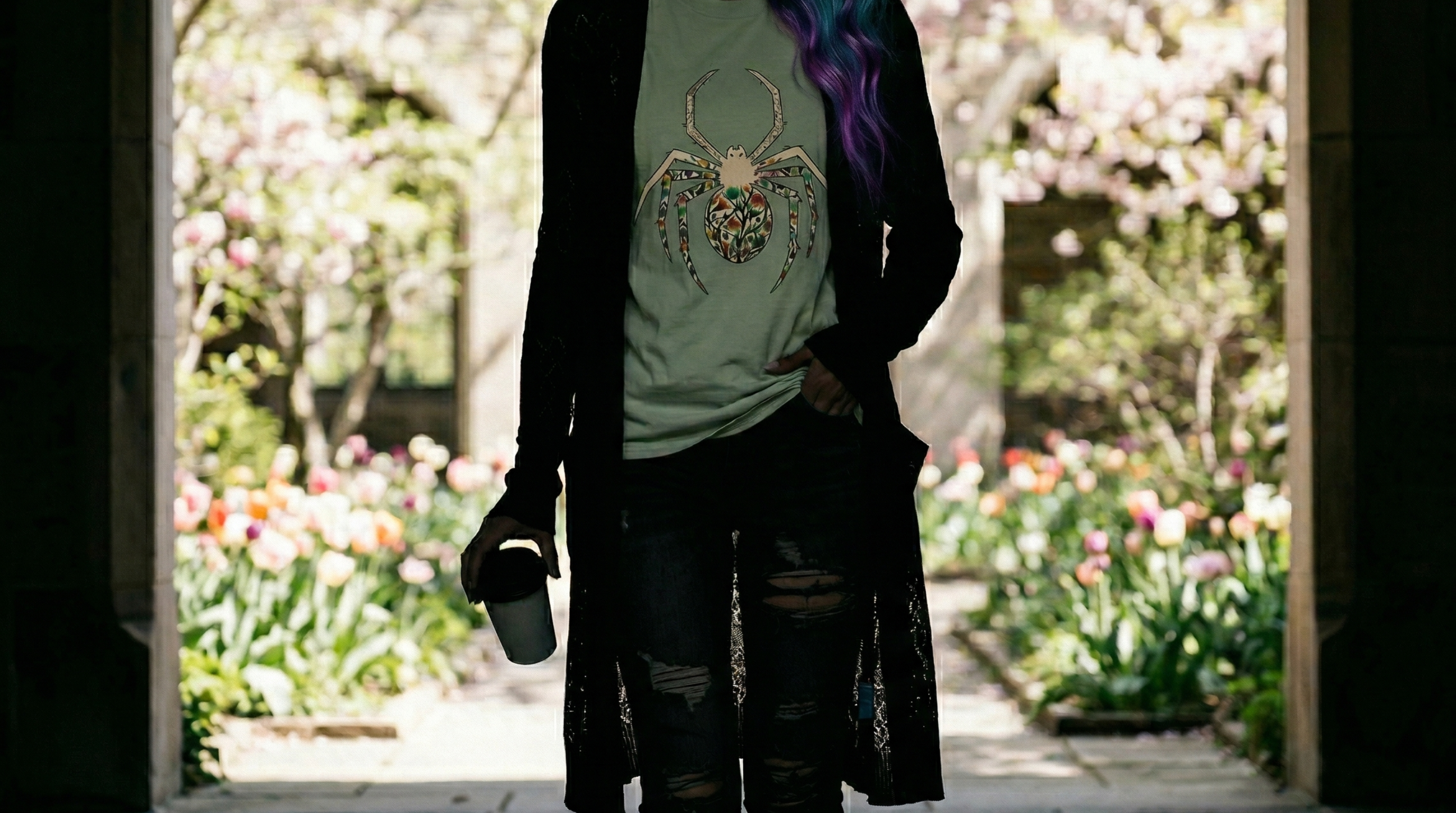 Closeup on a woman holding a coffee cup wearing a floral spider t-shirt under a black lacy cardigan in a moody alcove with a spring garden behind her