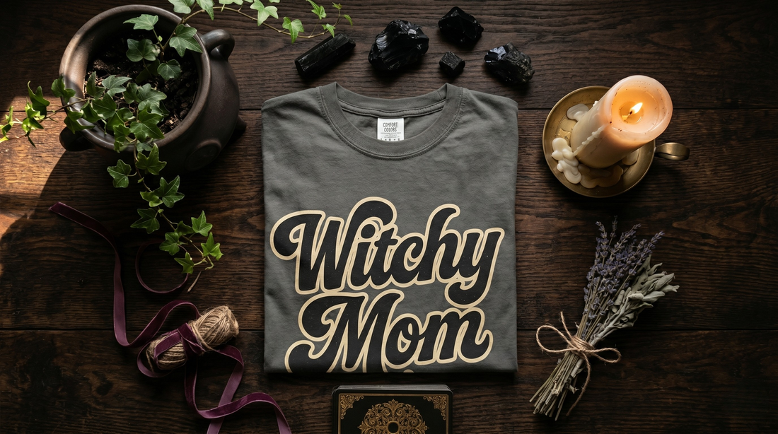Dark moody overhead flat lay on a textured dark wood surface. A folded Pepper Grey t-shirt ablazed with "Witchy Mom" in centered surrounded by witchy ephemera and accessories. 